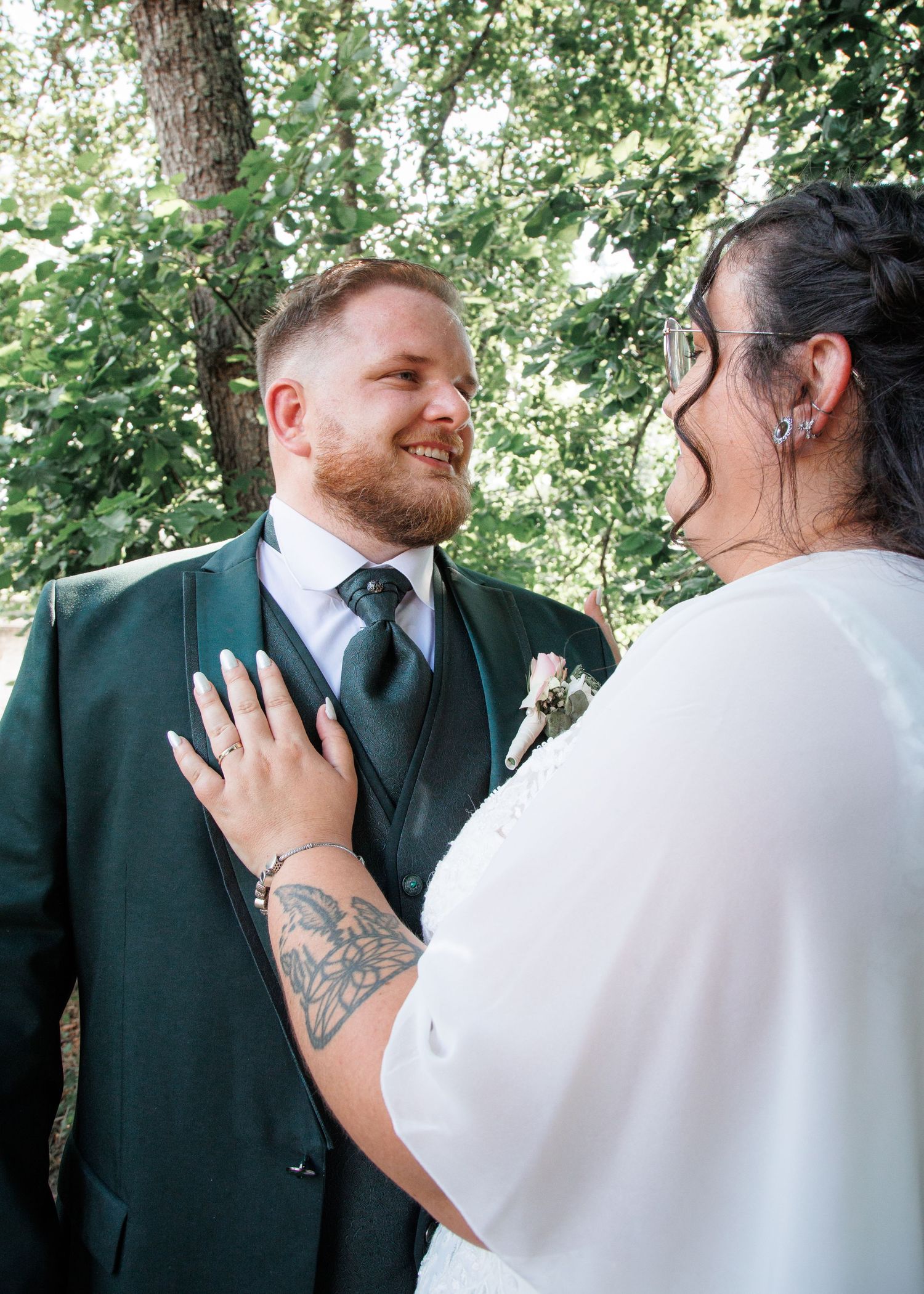 Bride and groom share a joyful moment outdoors, groom in green suit with boutonniere, bride in white dress with tattoos.