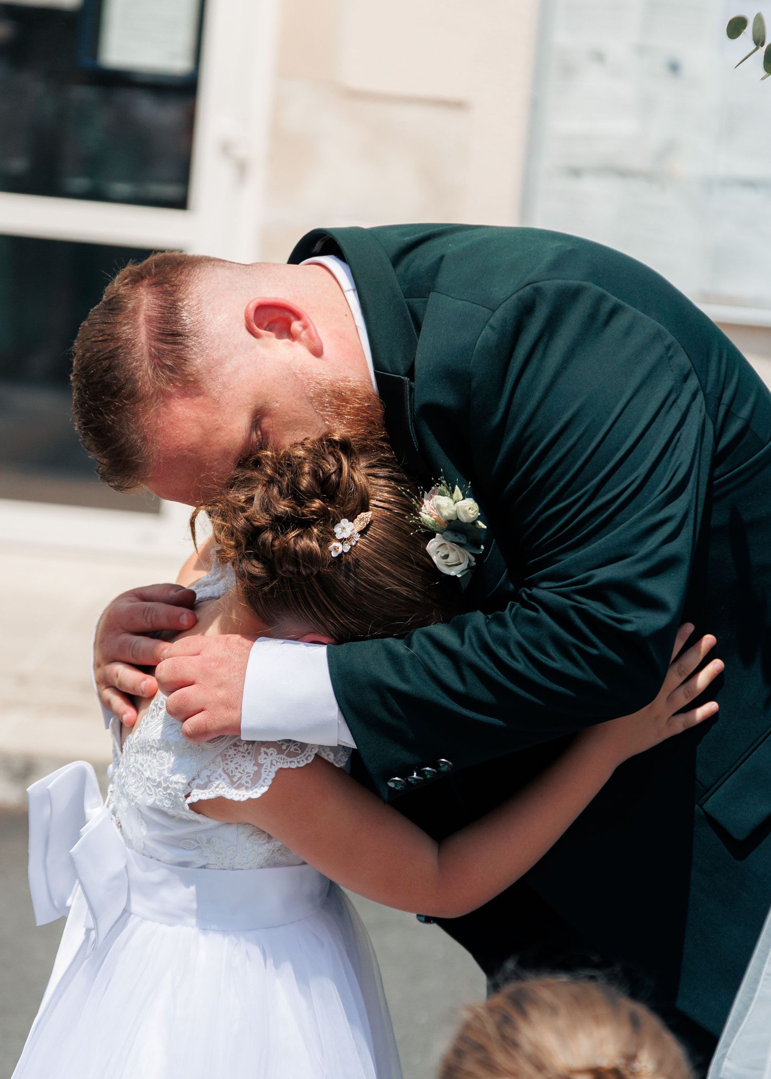 Emotional groom in dark green suit embracing a young flower girl in white dress at wedding ceremony.