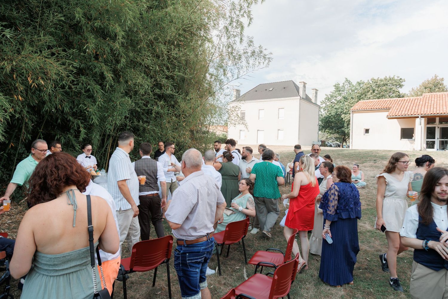 Wedding guests socializing outdoors in a garden near a French country house during summer reception.