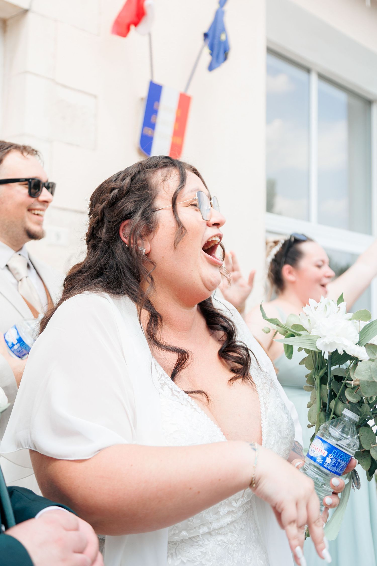 Bride in white dress laughing joyfully outside town hall decorated with French and EU flags.