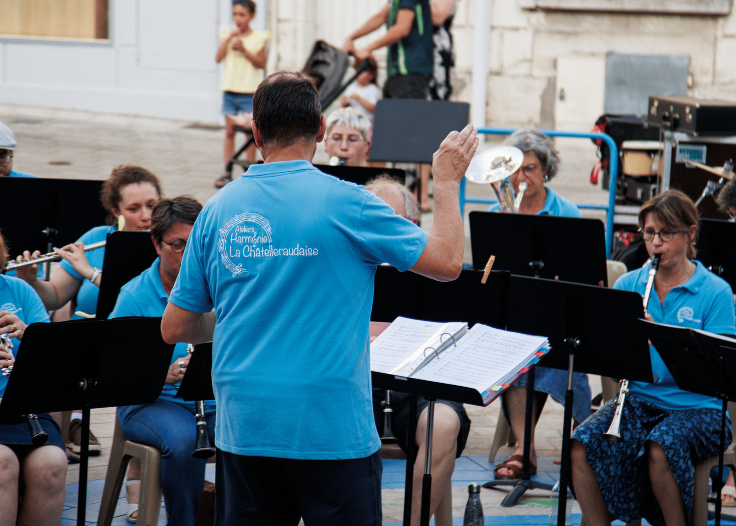 A conductor leads a band wearing bright blue shirts during an outdoor performance.