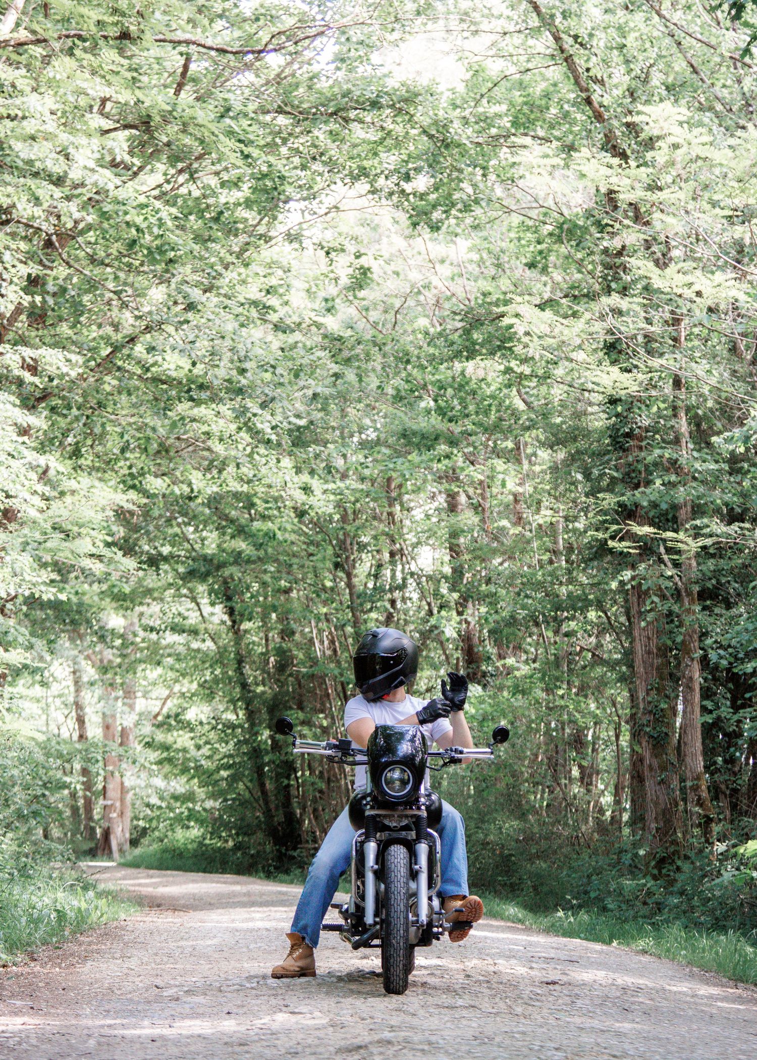 A motorcyclist in a helmet and jeans rides down a scenic tree-lined dirt road surrounded by lush green forest.