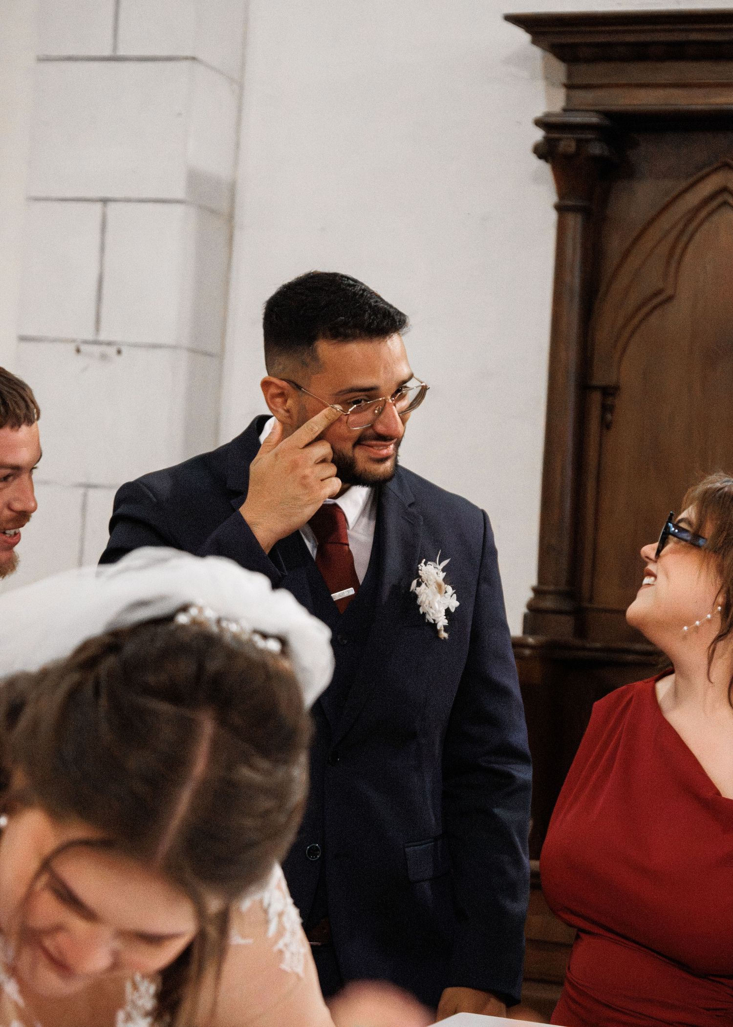 Emotional groom in navy suit wiping tears during wedding ceremony surrounded by guests in elegant venue.