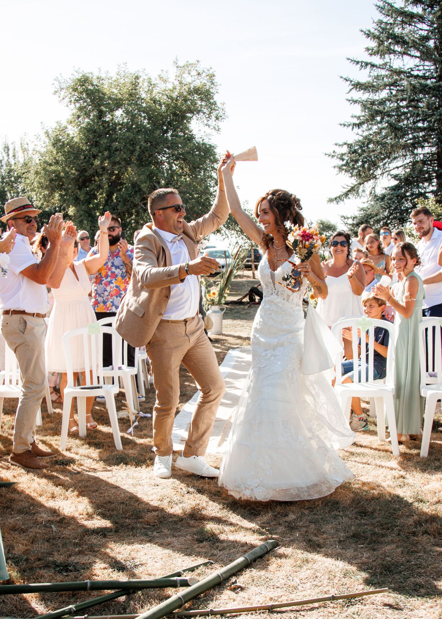 Wedding party celebrates outdoors by jumping in unison on a sunny afternoon in white attire with trees in background.
