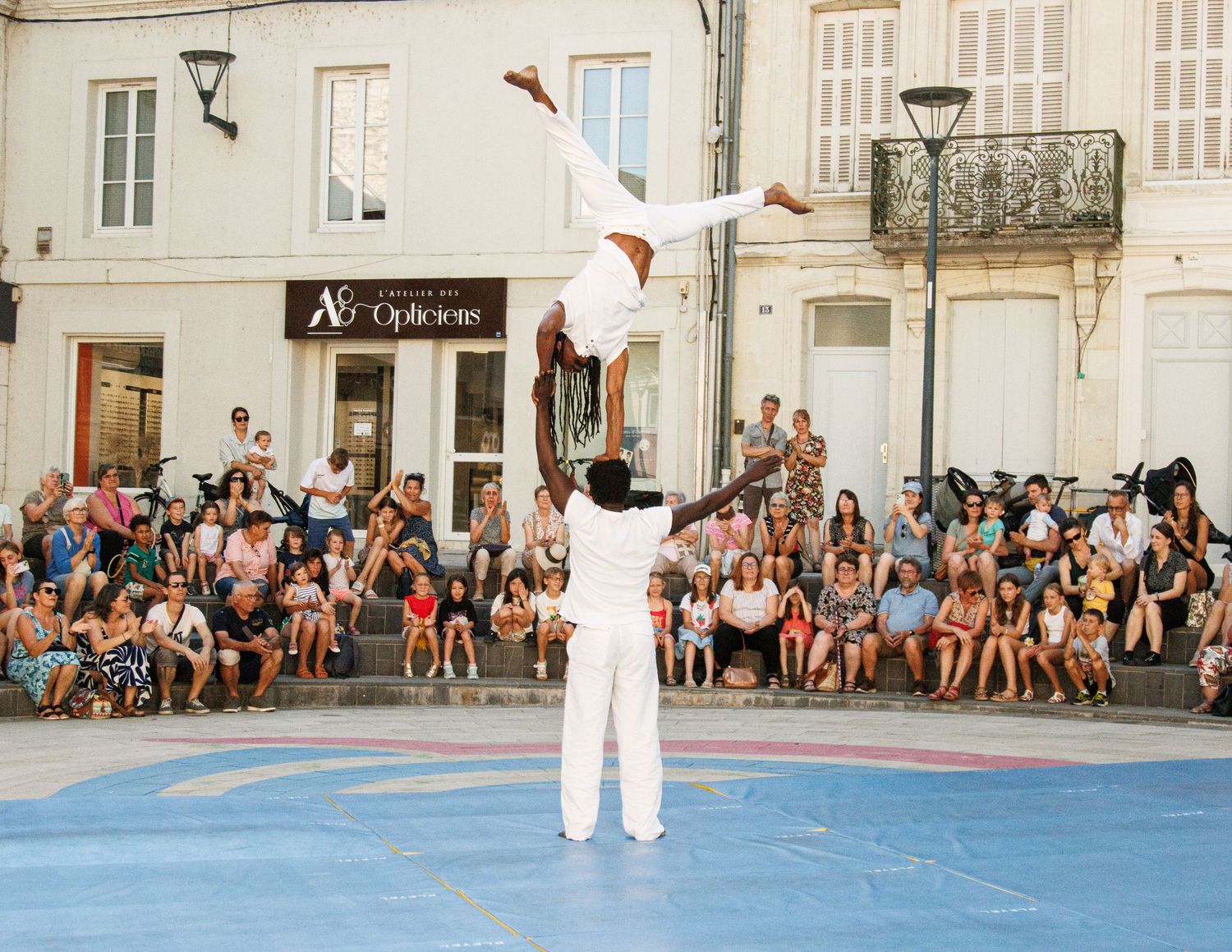 Street performers in white outfits demonstrate an impressive acrobatic balance routine on a blue performance mat.