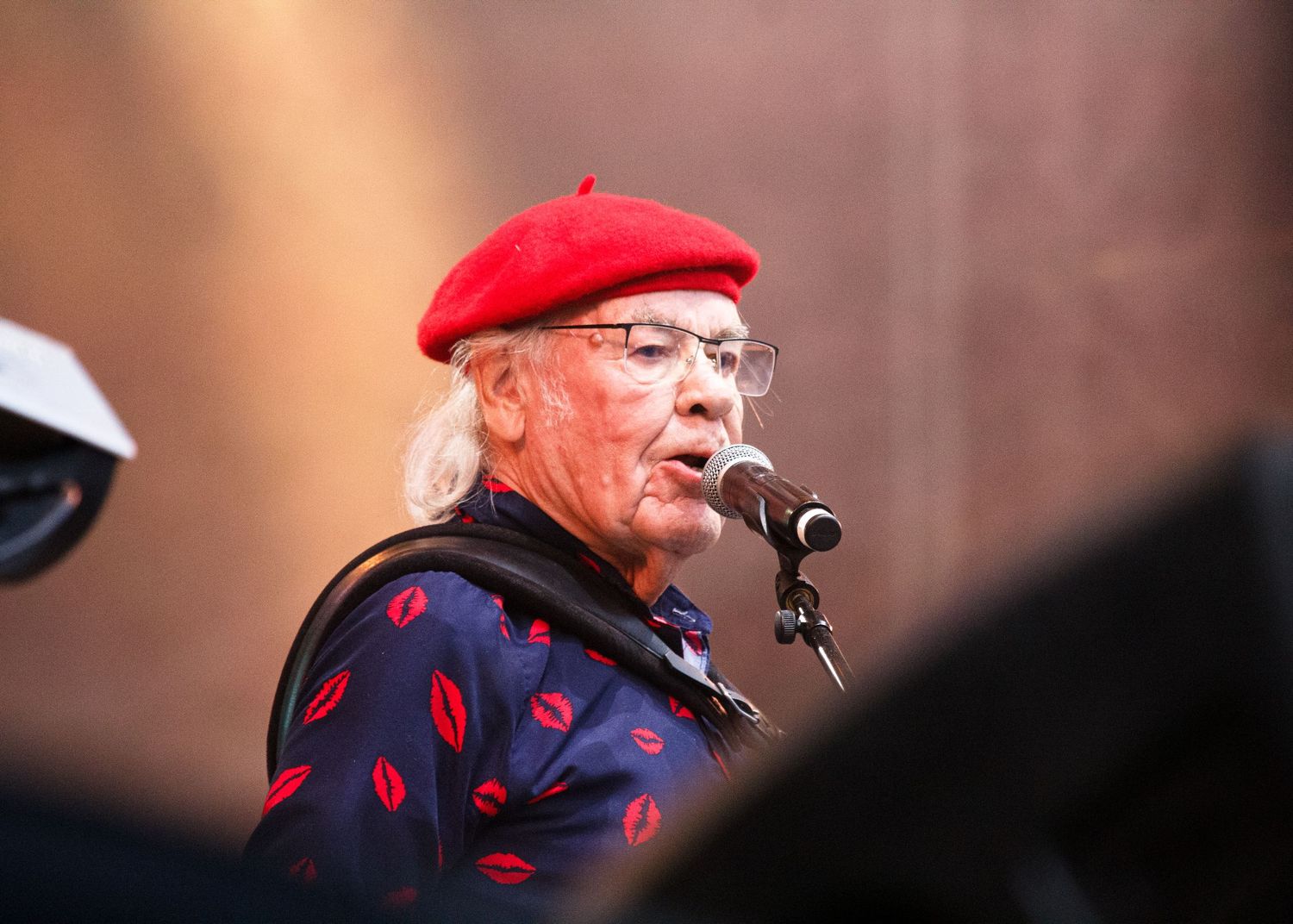 Someone wearing glasses and a red beret smokes a pipe while wearing a black and red patterned shirt outdoors.