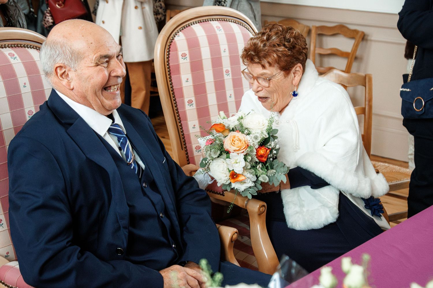 Elderly couple smiling at a wedding, man in navy suit, woman in white jacket holding a bouquet of flowers.