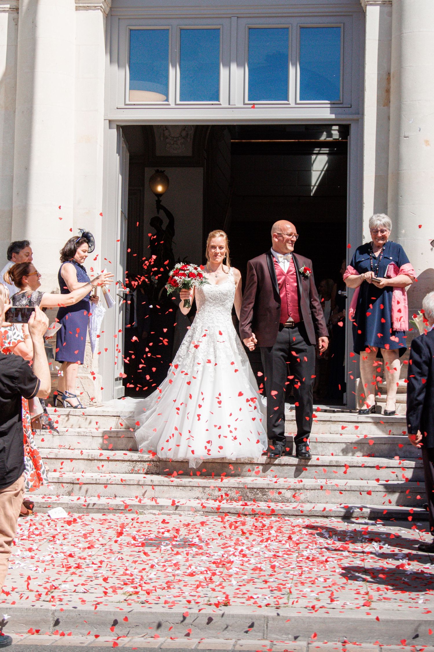 Newlyweds exit church steps as guests shower them with red rose petals, bride holding red bouquet in white gown.