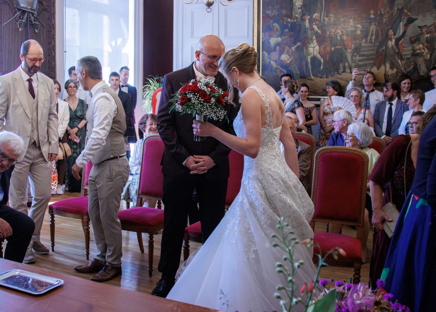 Bride in lace wedding gown exchanges bouquet with elderly man during indoor ceremony.
