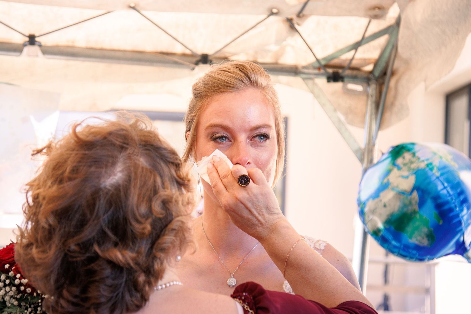 Woman carefully applies makeup to a young blonde bride at an outdoor wedding celebration under a tent.