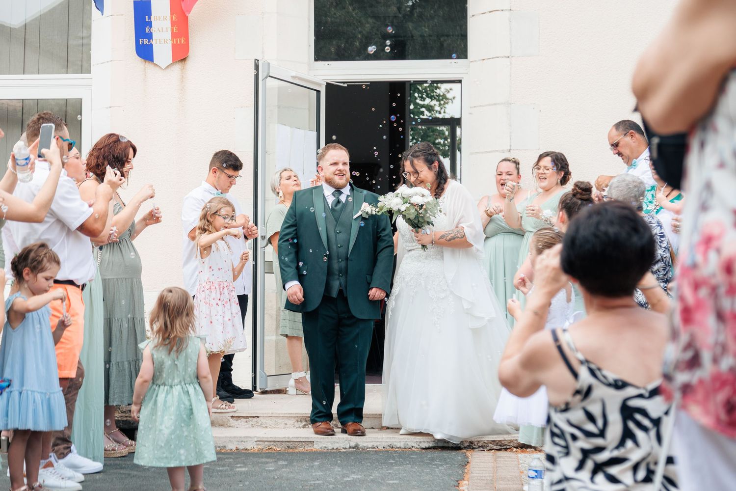 Newlyweds exit ceremony hall as guests in sage green attire celebrate with confetti on their wedding day.