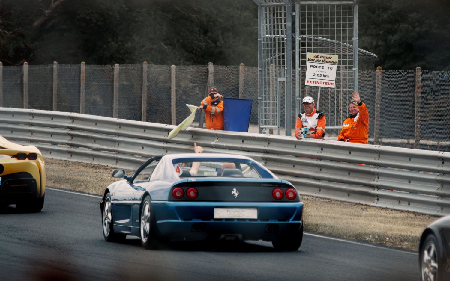 Blue sports car racing on track with safety barriers and race marshals in orange uniforms.