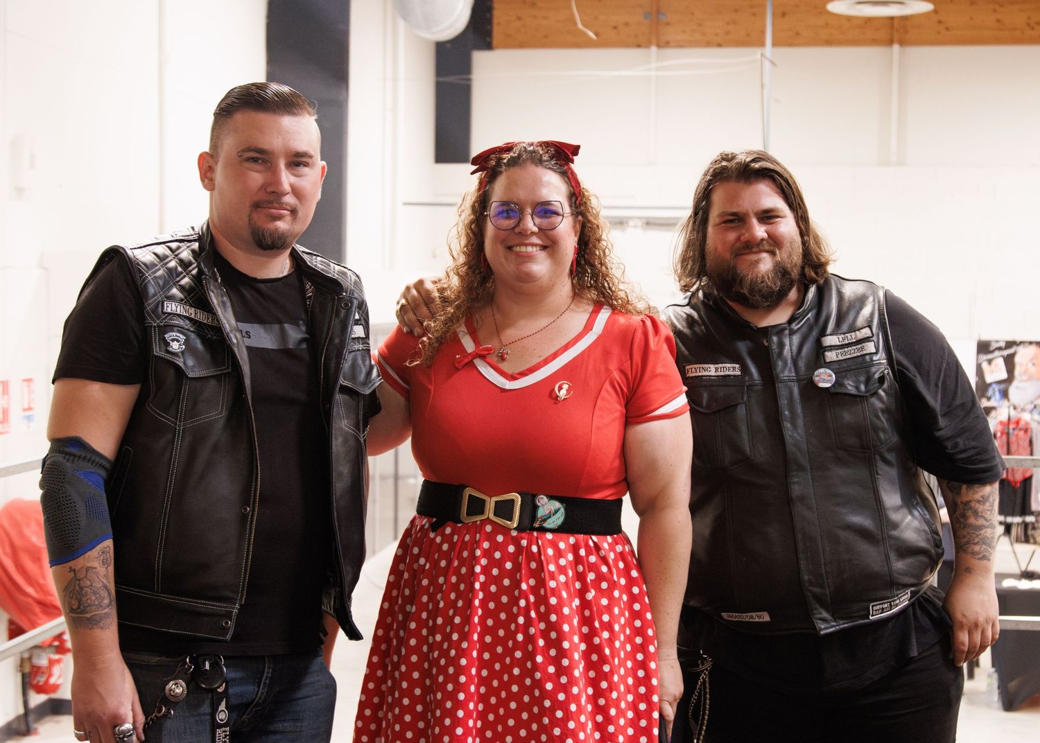 People in leather vests stand together with someone in a red polka dot dress.