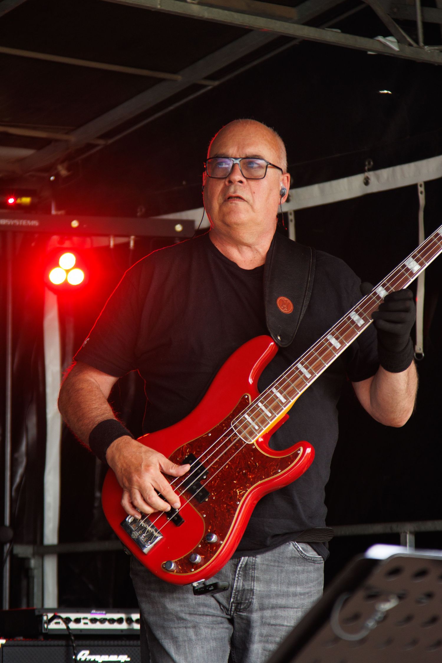 Bass guitarist performs on stage with a bright red electric bass guitar under red stage lighting.
