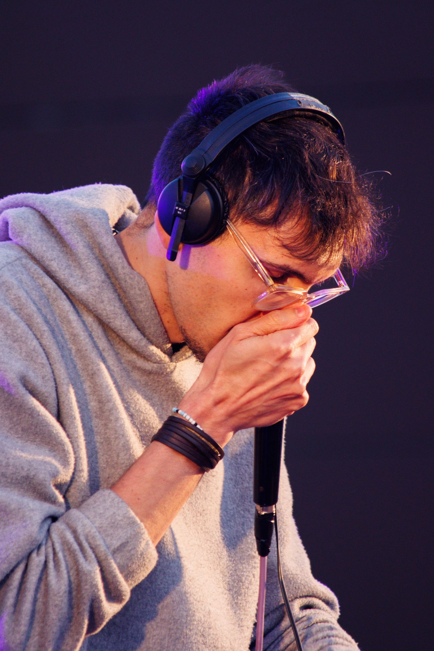 Performer in gray hoodie wearing headphones while singing into microphone on dark stage with purple lighting.