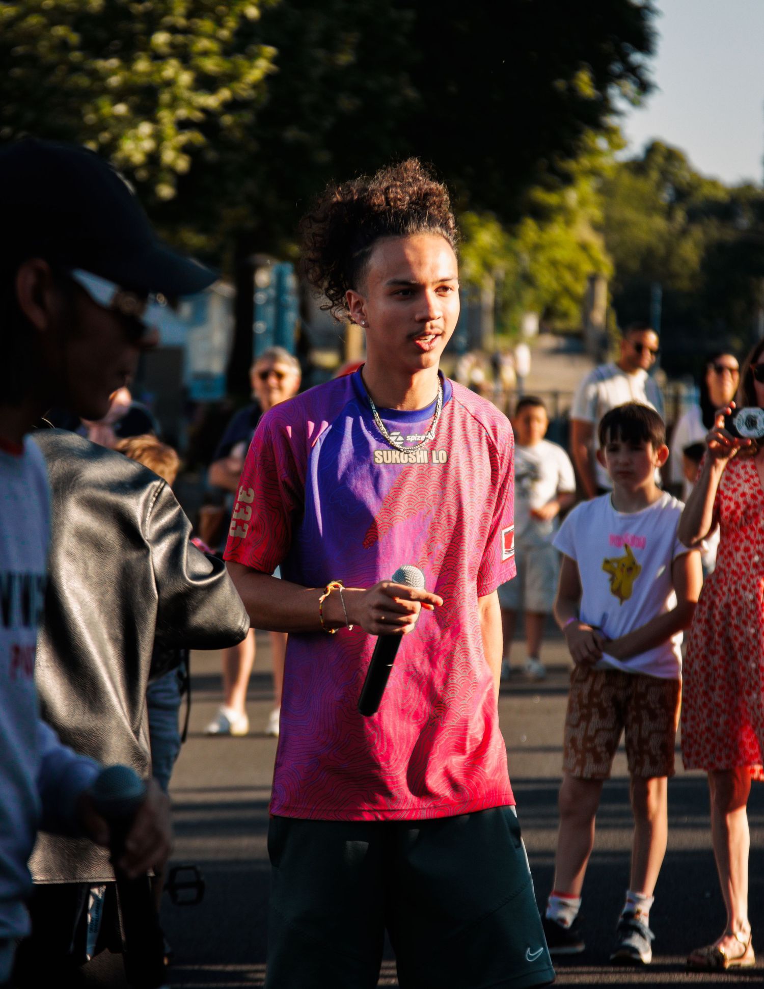Performer in pink and purple t-shirt stands outdoors among crowd during daytime event.