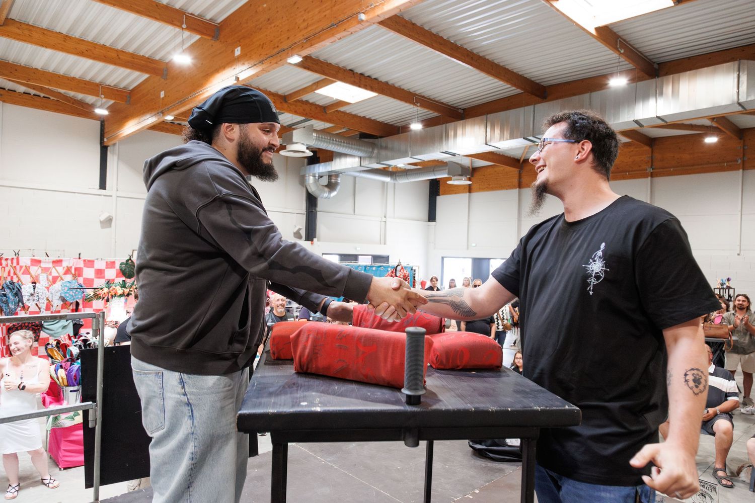 Two men shaking hands at an arm wrestling competition table in a packed indoor sports venue.