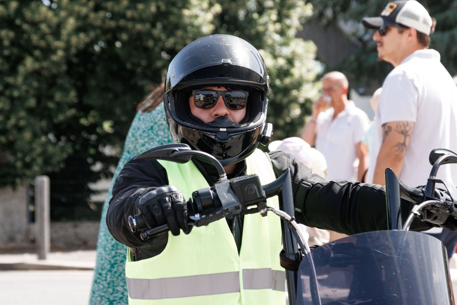 Motorcyclist wearing a black helmet, sunglasses, and green safety vest adjusts mirrors on a motorcycle outdoors.