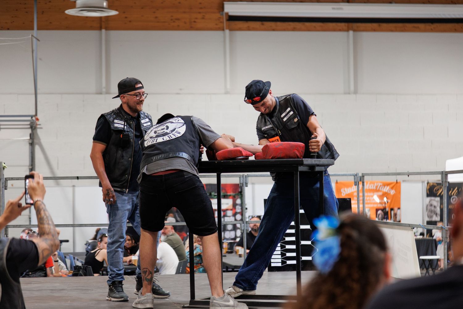 Two men compete in an arm wrestling match on a red padded table at an indoor sporting event.
