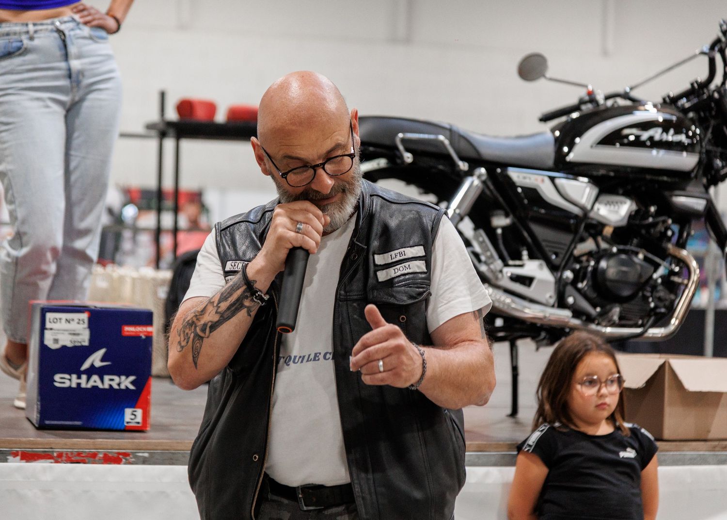 Bald tattooed man in biker vest speaks into microphone at motorcycle event with young girl and bike in background.