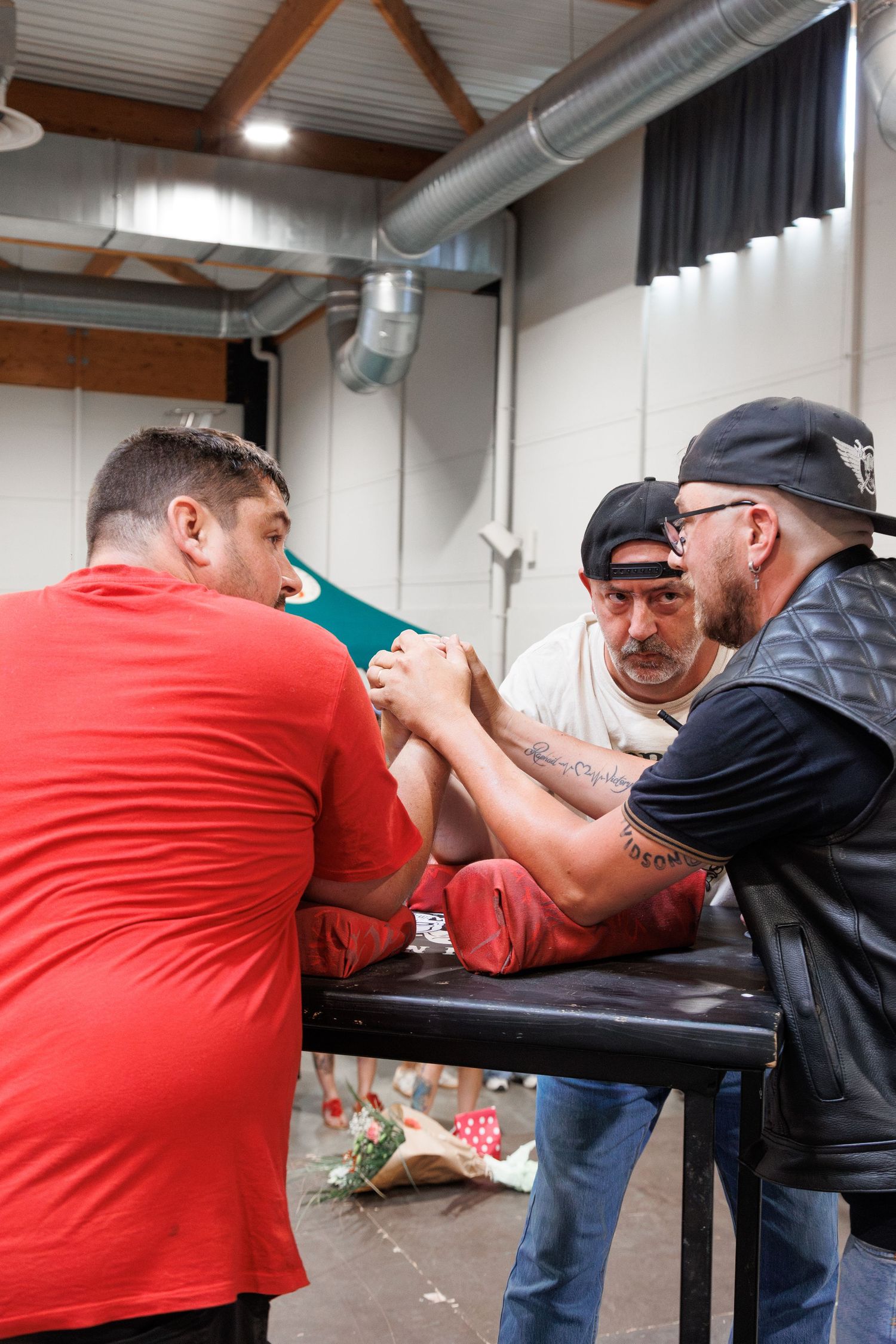 Two men arm wrestling at a table while a referee watches closely in an indoor venue.