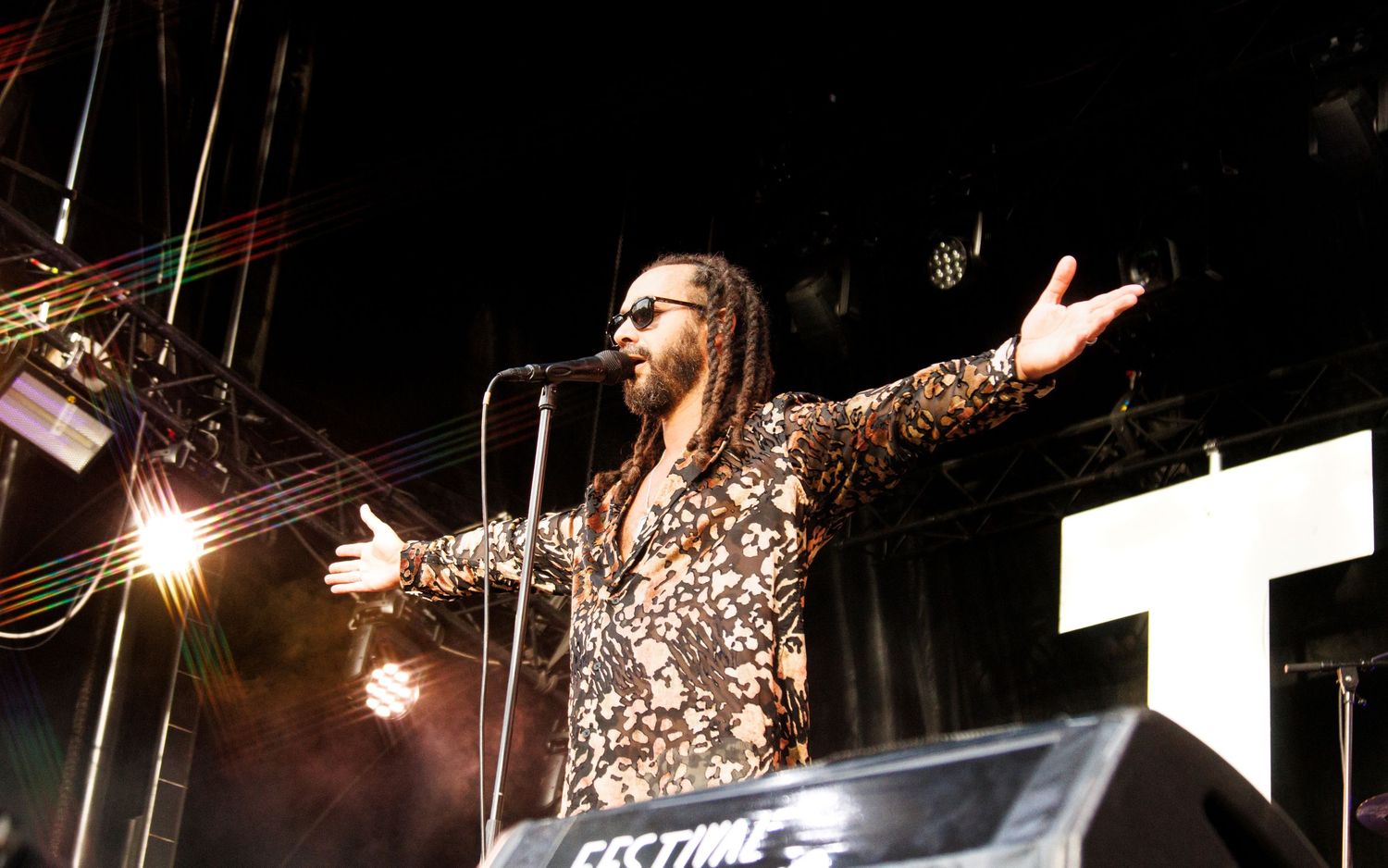 A performer with dreadlocks in a floral suit sings on stage with arms outstretched at an outdoor music festival.