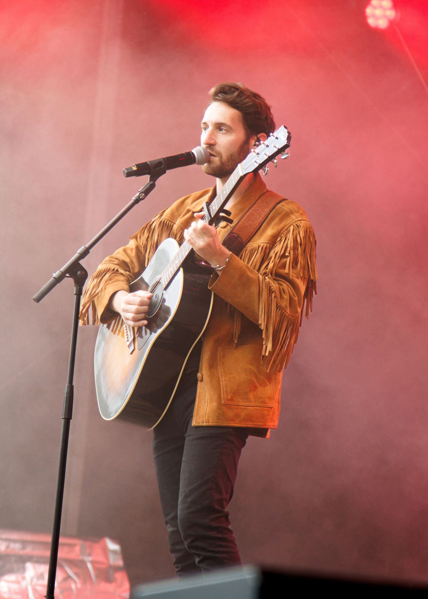 A performer in a brown fringed jacket plays acoustic guitar on stage bathed in dramatic red lighting.