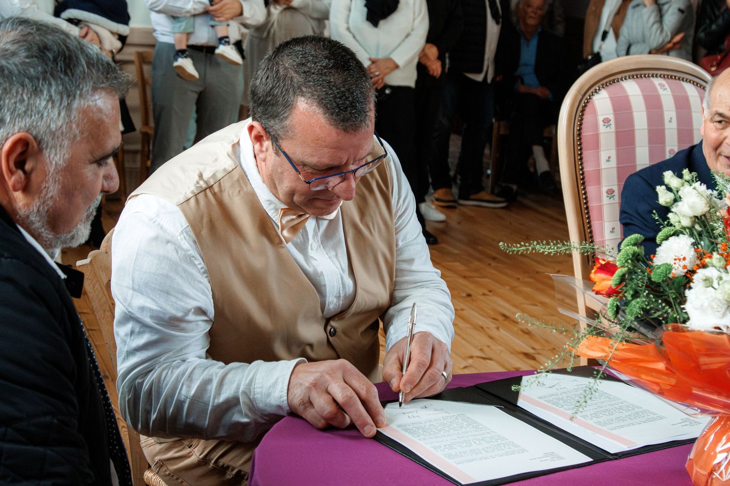 Man signing official documents at a formal ceremony table with attendees gathered around in an elegant room.