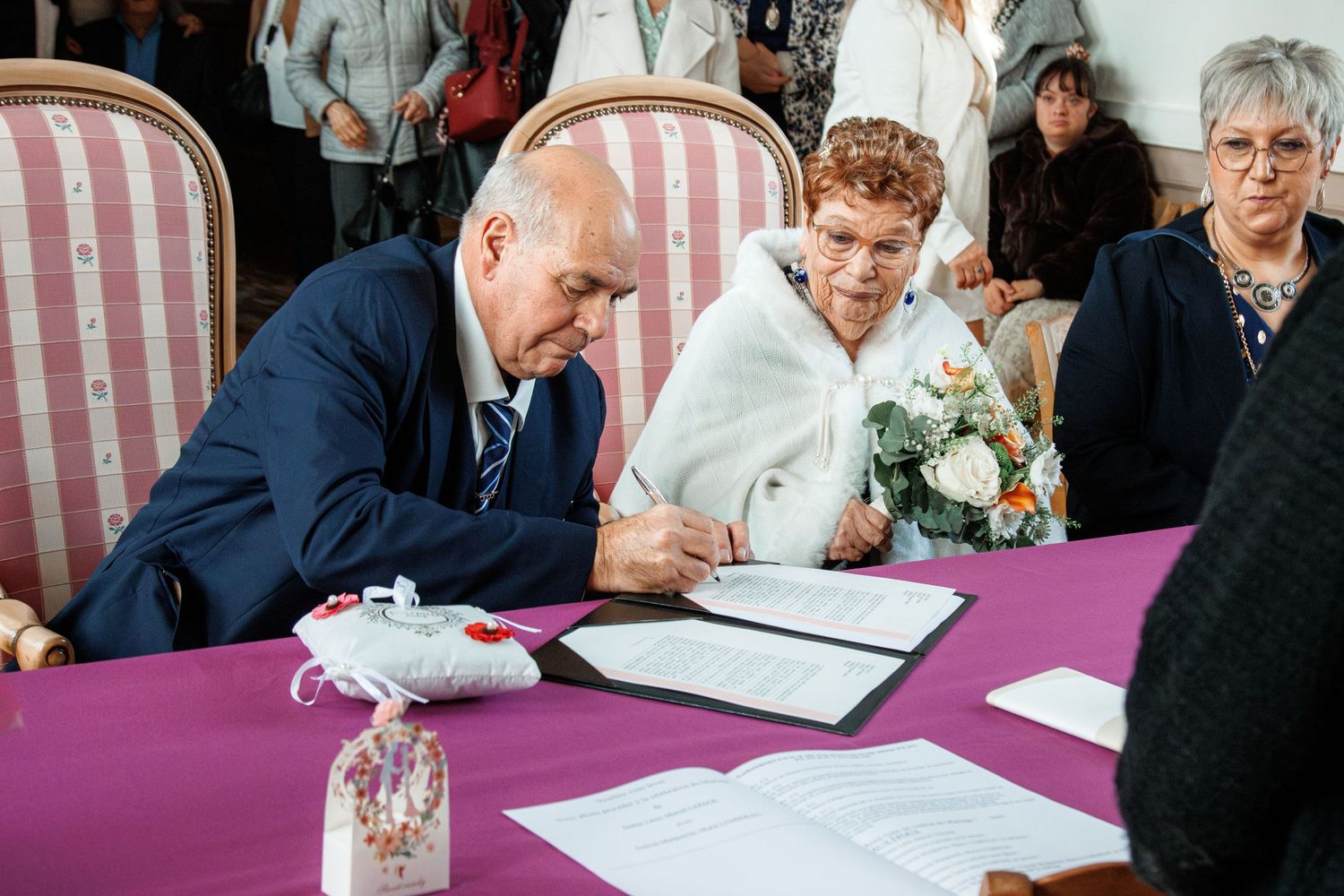 Elderly couple signing marriage documents at a purple-draped table during a civil wedding ceremony.