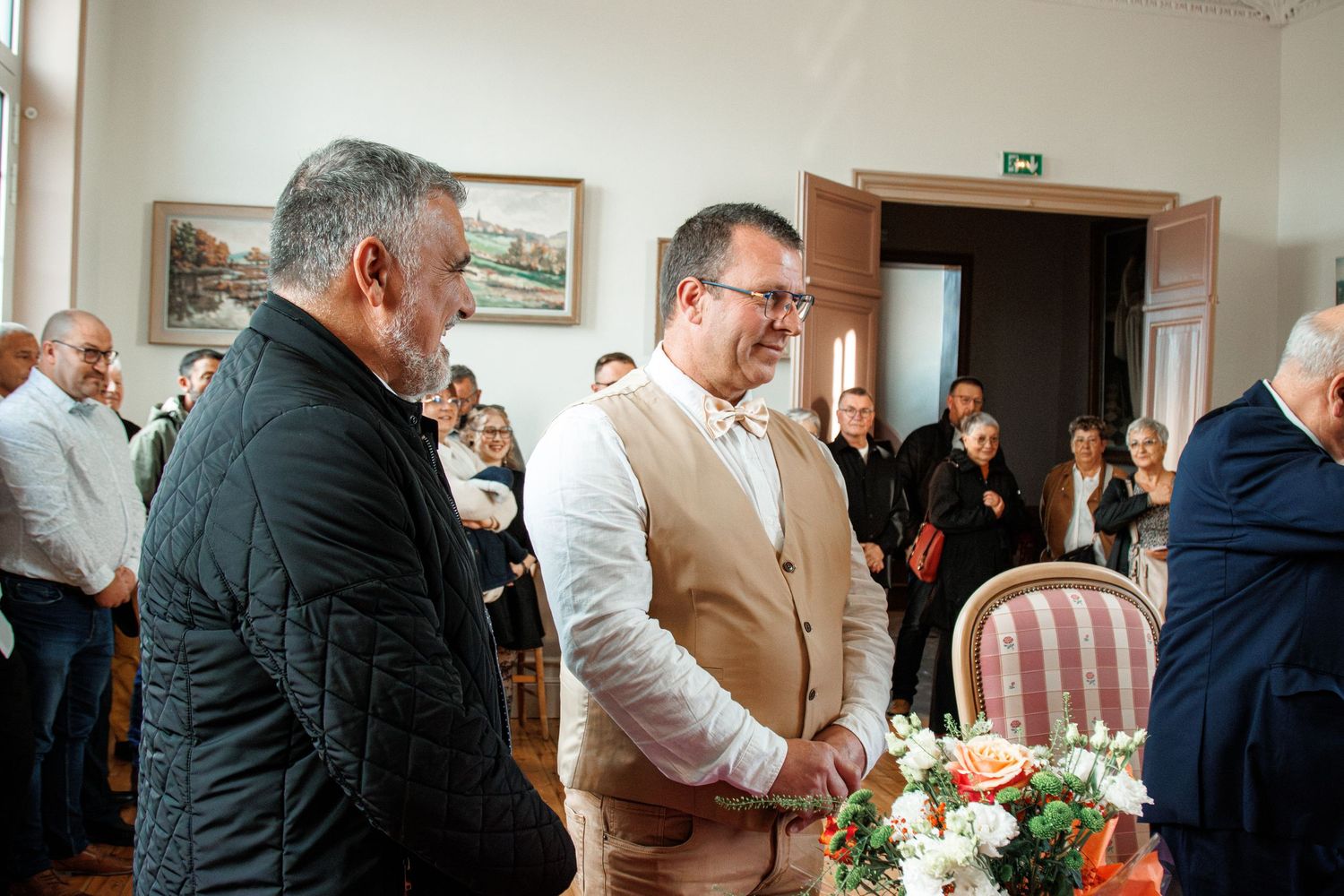 Two men shaking hands at a formal indoor gathering with guests in the background.