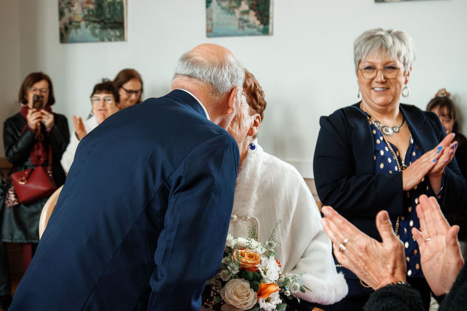 Elderly couple sharing a kiss at a wedding ceremony while guests applaud and smile in the background.