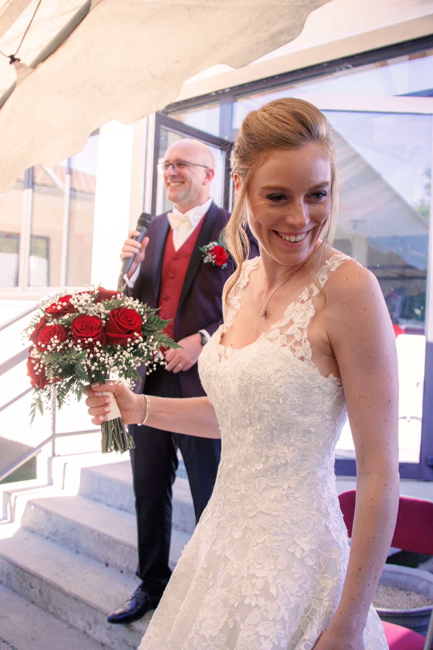 Radiant bride in lace gown holding red bouquet smiling on steps with groom behind her.
