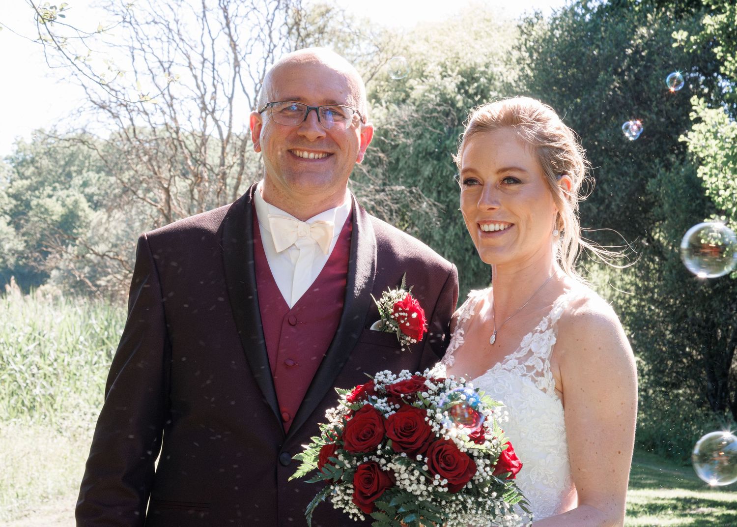 Smiling bride and groom close-up portrait outdoors, bride holding red rose bouquet with soap bubbles floating nearby.