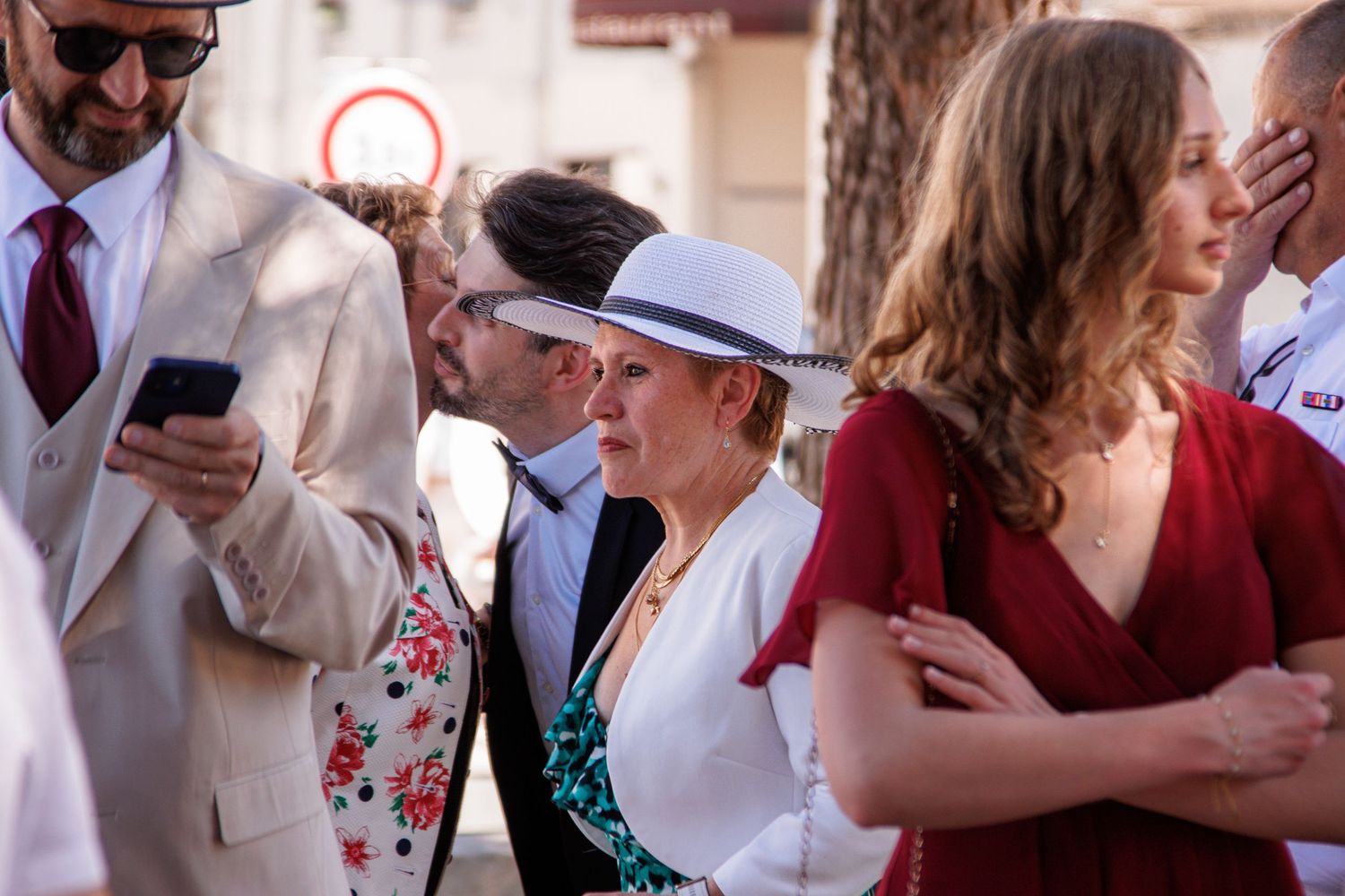 Elegantly dressed guests at an outdoor event, woman in white hat and jacket checks phone on sunny day.