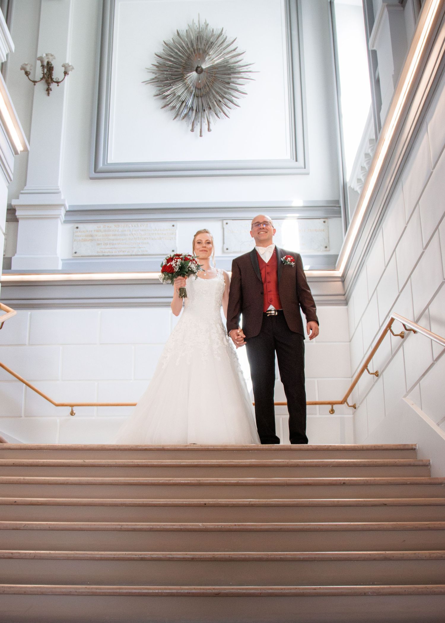 Bride and groom holding hands on grand staircase, bride in white lace gown holding red bouquet, groom in dark suit with red vest.