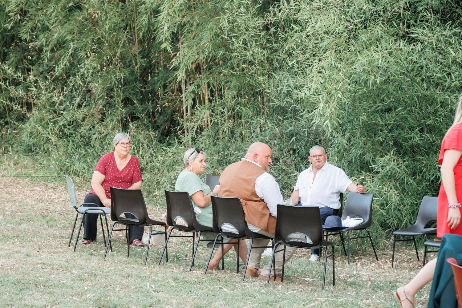 Four wedding guests seated on black chairs conversing outdoors near green bamboo trees.