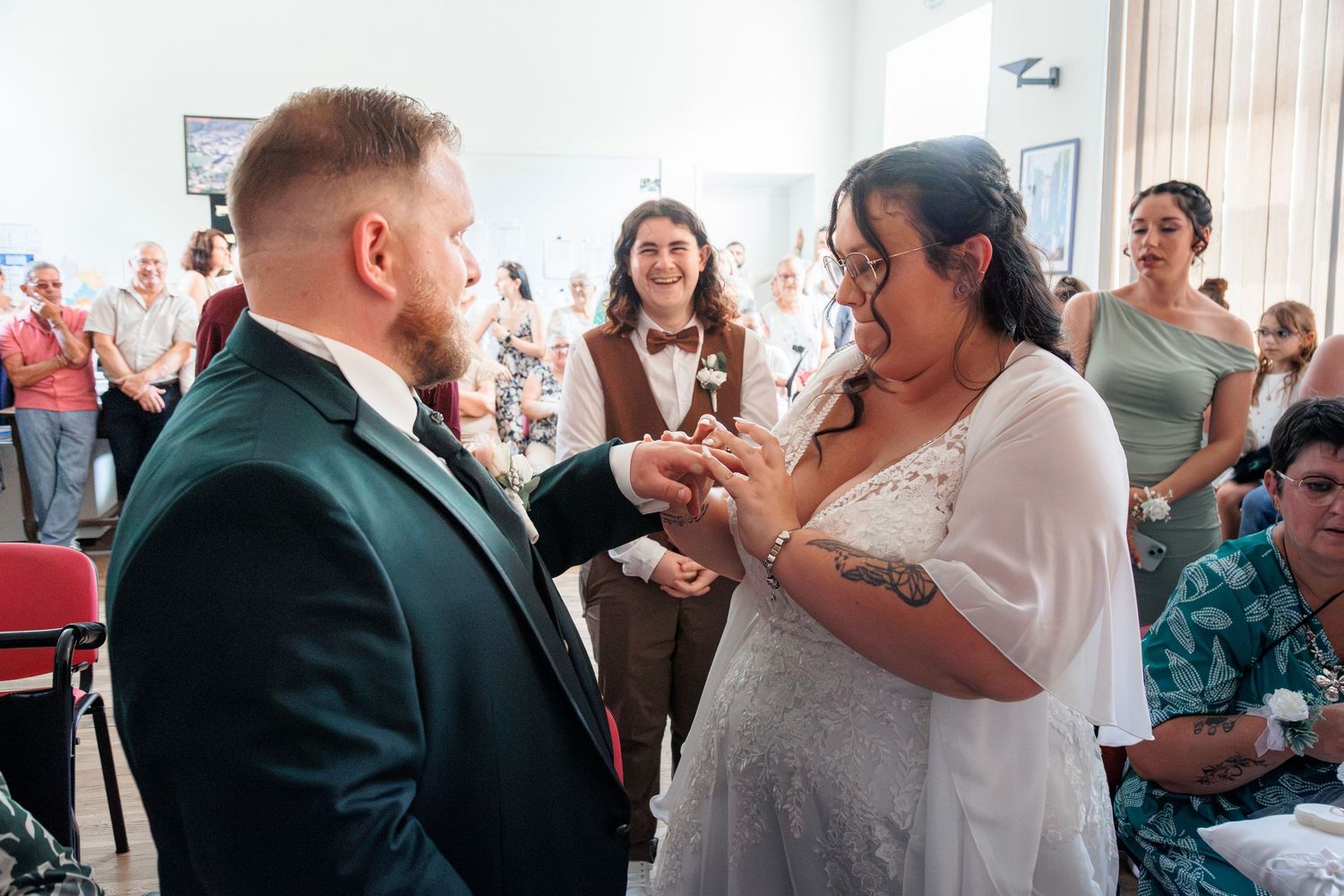 Bride placing wedding ring on groom's finger during indoor ceremony with guests watching in background.