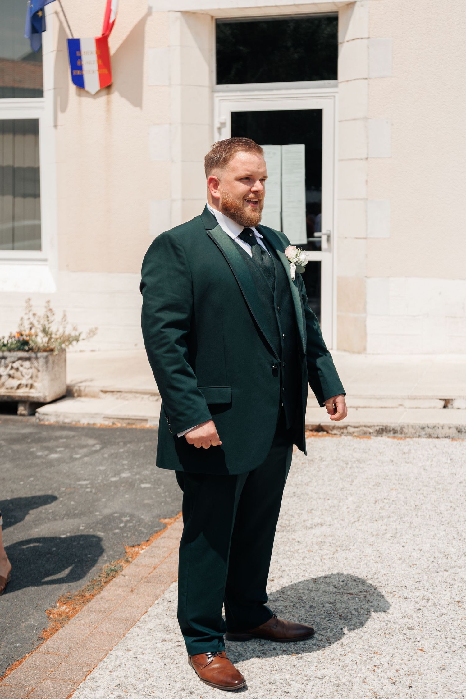 Smiling groom in elegant dark green suit with boutonniere stands outside a French mairie building.