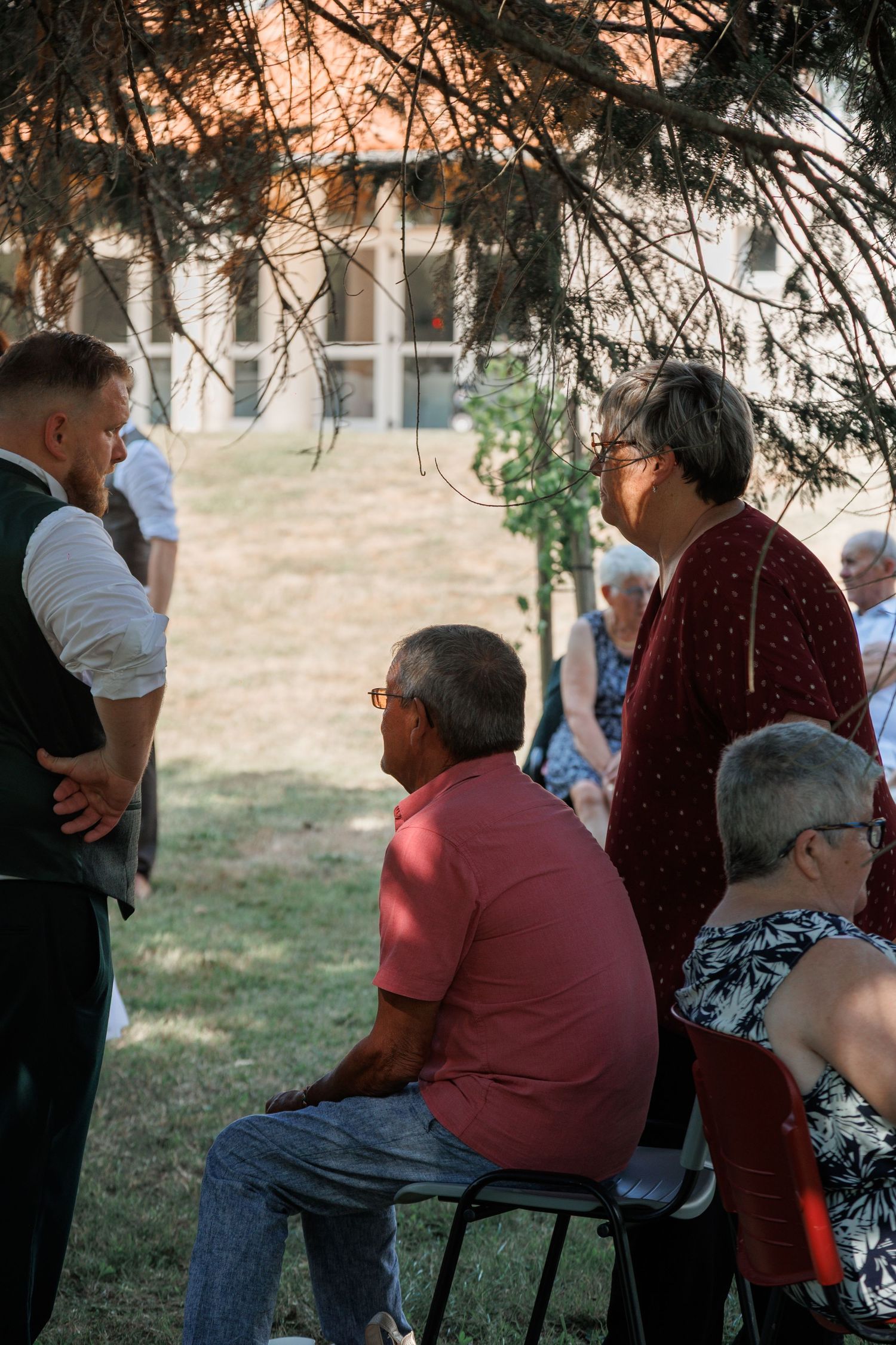 Wedding guests seated outdoors under tree shade watching ceremony in sunlit garden setting.