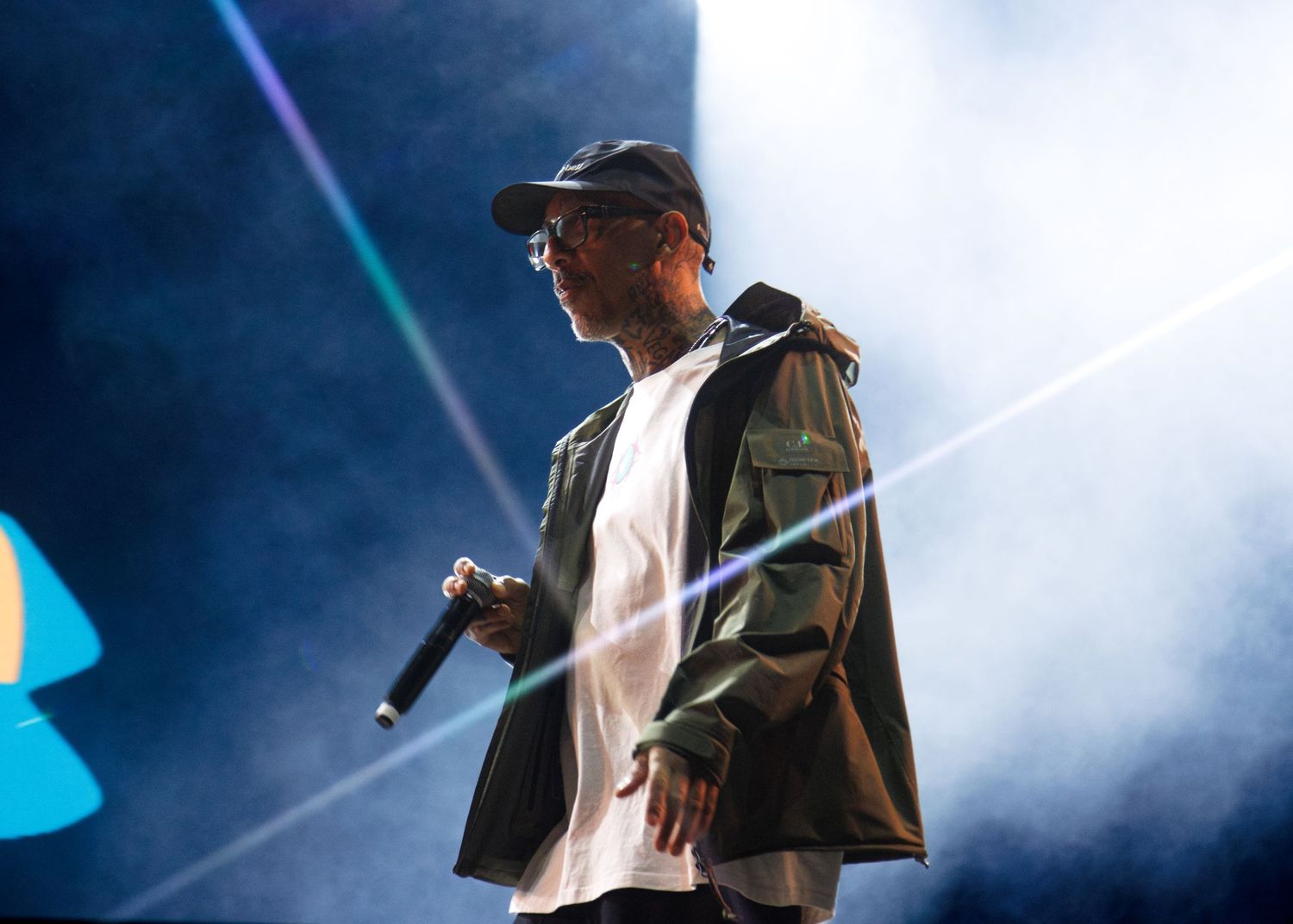 Performer on stage holding microphone in olive jacket and white shirt with dramatic concert lighting and fog effects.