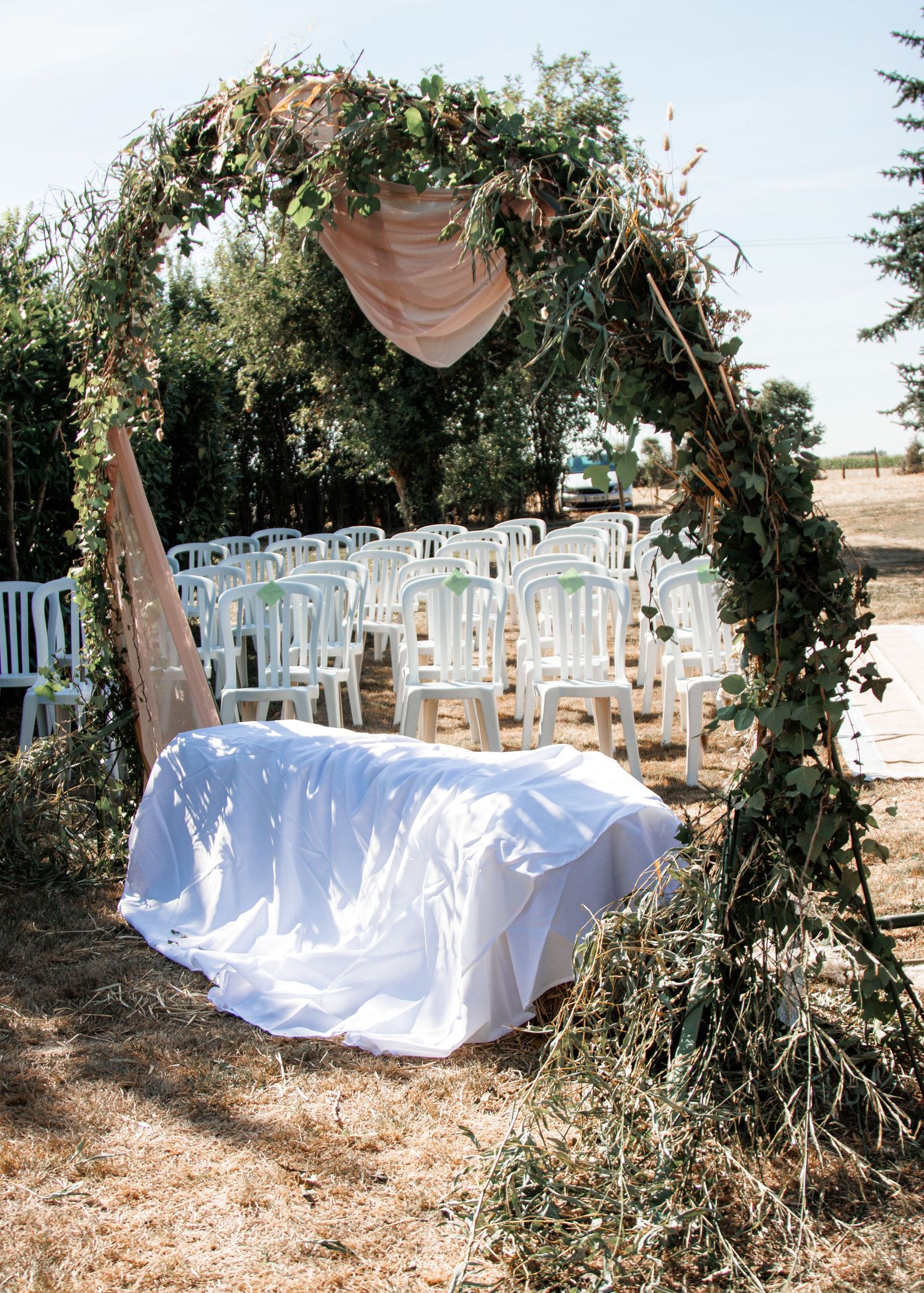 A rustic outdoor wedding ceremony setup with white chairs and a natural wood arch decorated with greenery and white fabric.
