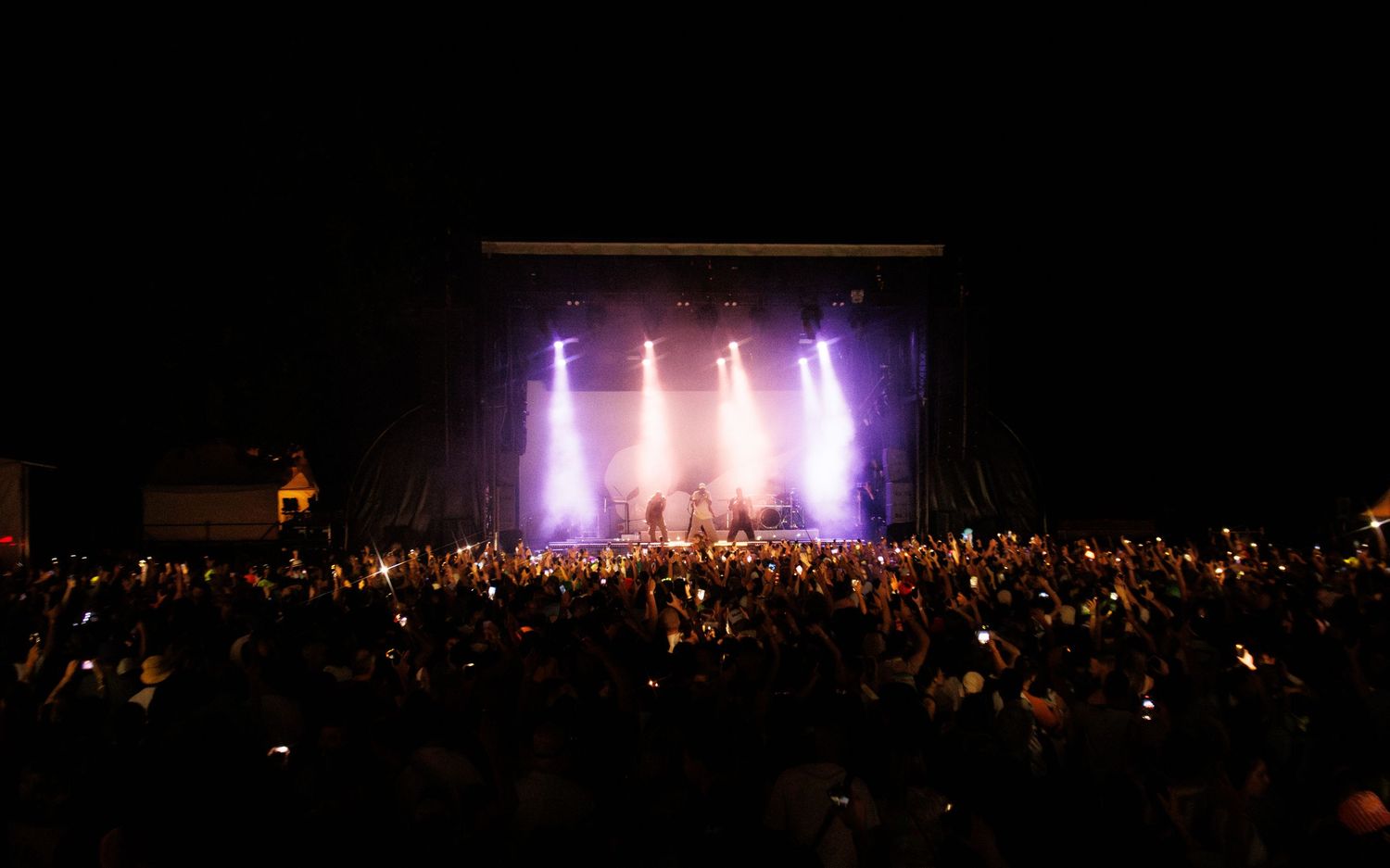 Concert crowd silhouettes stand before illuminated stage lights during an outdoor nighttime performance.