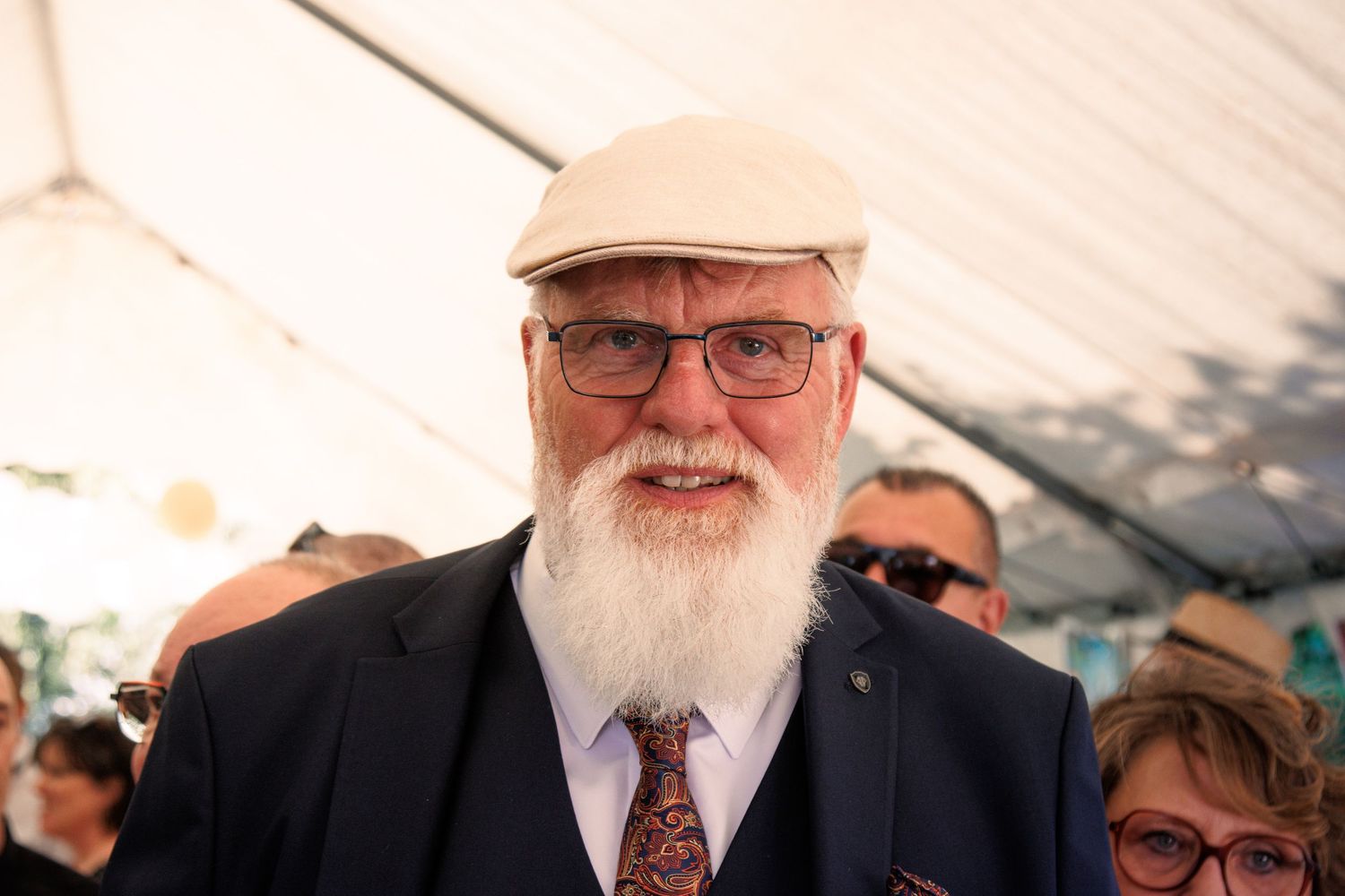 Cheerful elderly man with white beard wearing flat cap and glasses at wedding reception.