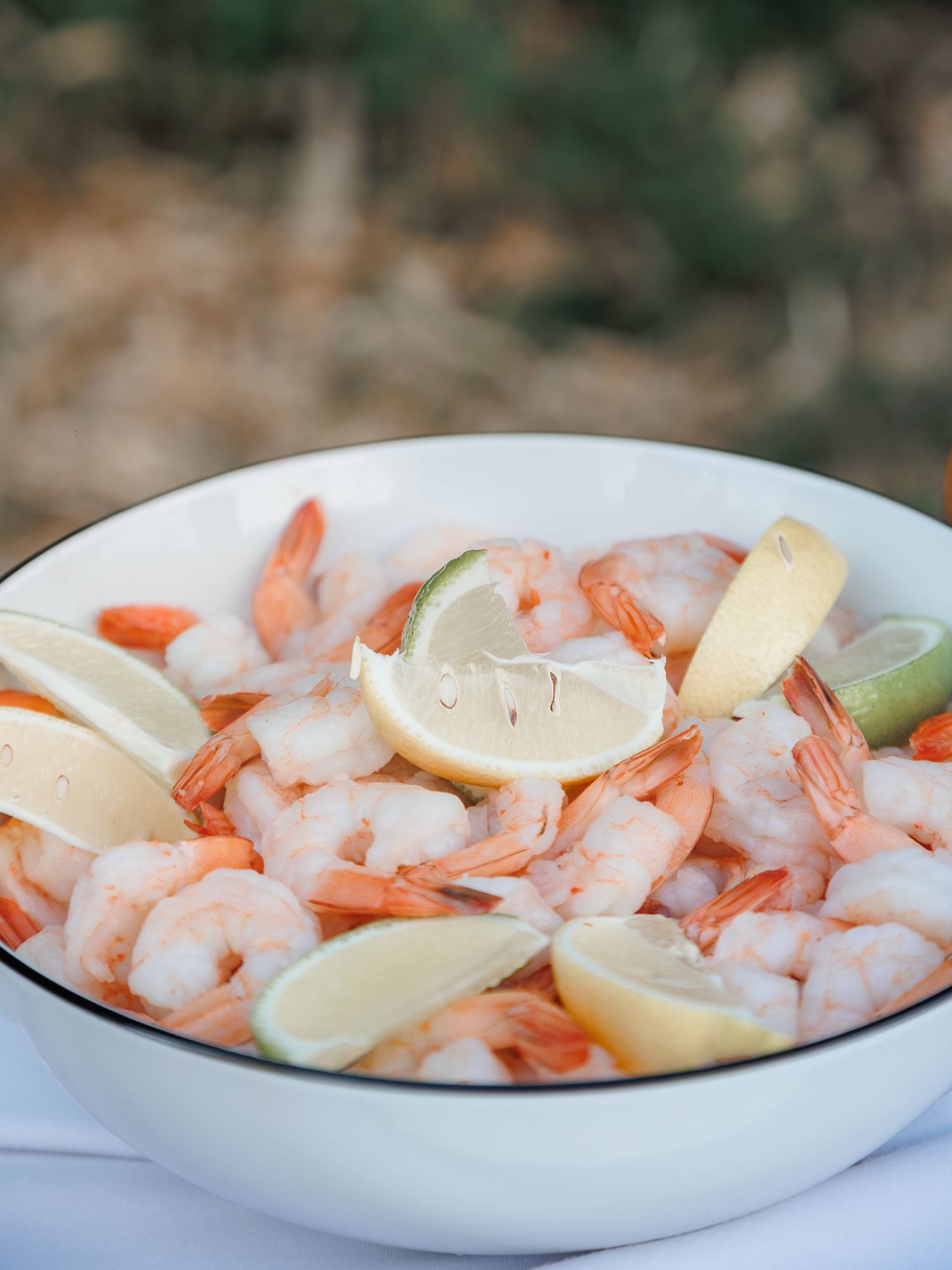 Large white bowl filled with fresh cooked shrimp garnished with lemon and lime wedges.