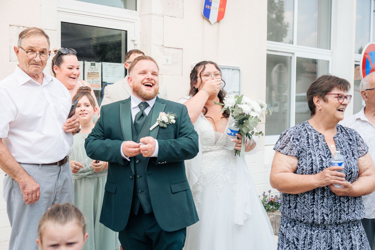 Laughing groom in green suit and bride with bouquet celebrate outside wedding venue with guests.