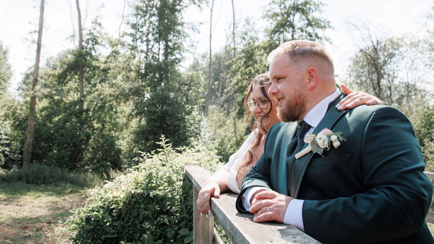 Bride and groom in green suit leaning on wooden fence surrounded by lush forest trees.