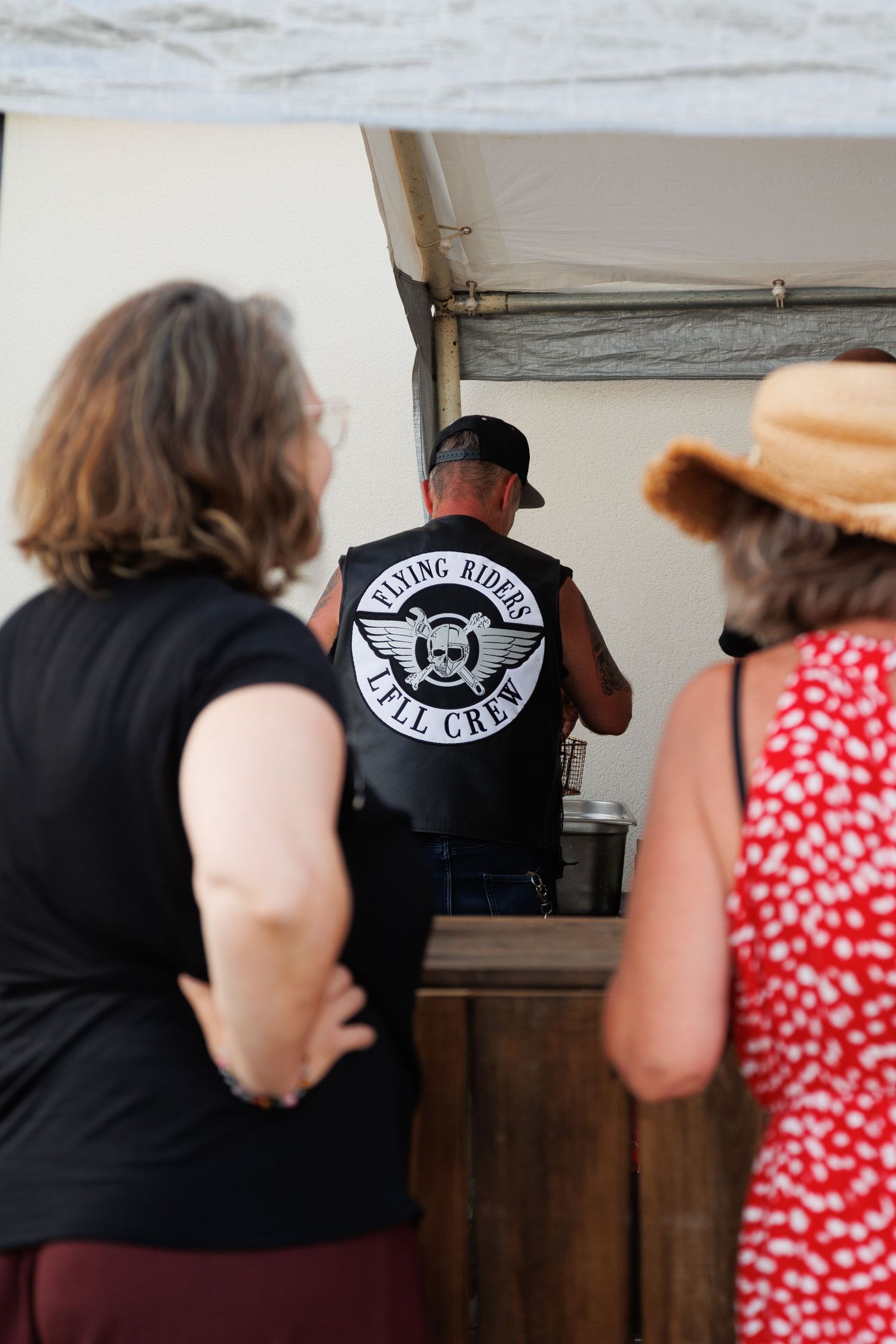 Man wearing Flying Riders Till Crew biker vest stands at outdoor food stand with two women waiting behind him.