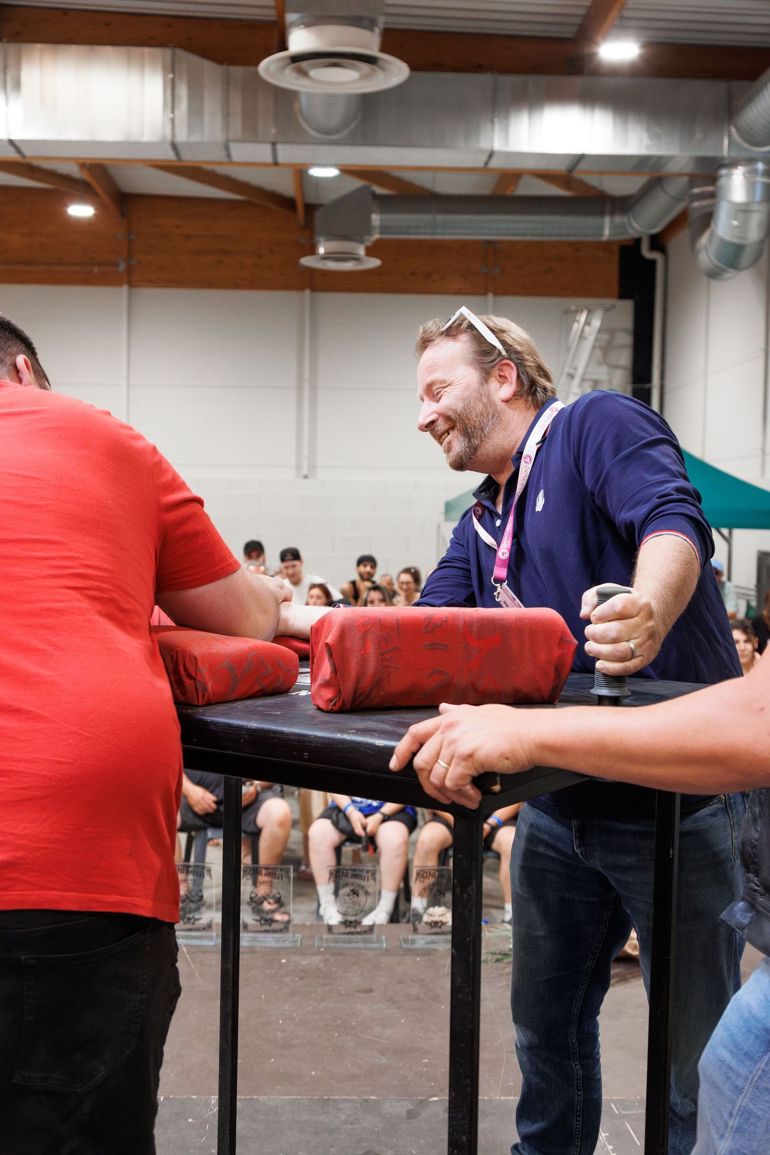 Two men compete in an arm wrestling match at an indoor event, with an audience watching in the background.