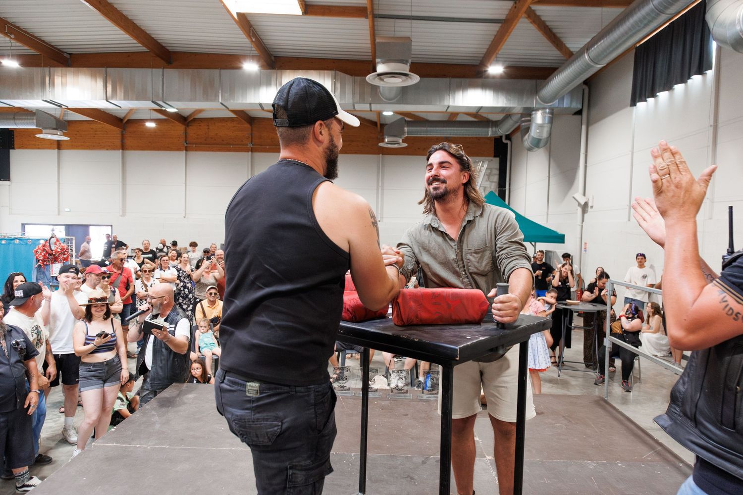 Two men arm wrestling at a table during an indoor competition event with a crowd of spectators watching.