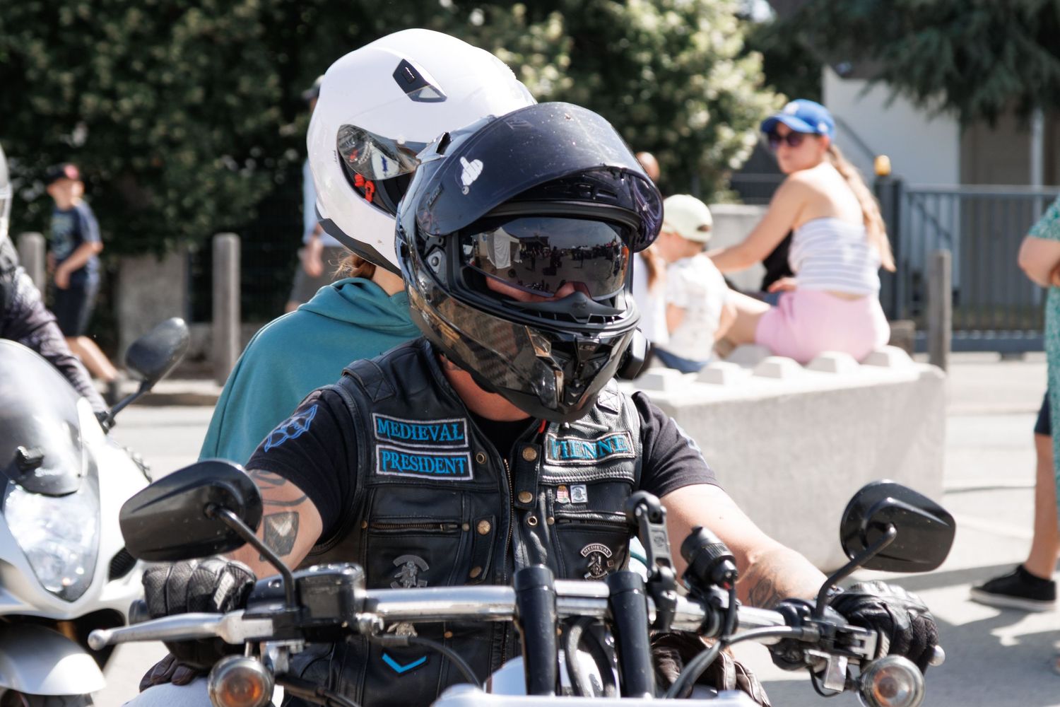 Two motorcyclists wearing helmets ride bikes at an outdoor event on a sunny day.